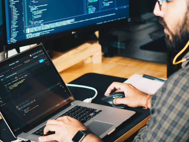 Bearded man wearing glasses and a plaid shirt working at a desk with a laptop and a large LG monitor displaying code in a dark-themed IDE