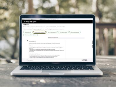 Laptop computer on a wooden surface displaying the Sage Help Agent interface in a desktop browser window, with suggested topic buttons and a response to a transaction reversal query visible on screen.