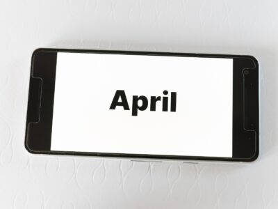 A smartphone lying horizontally on a white textured surface, screen facing up and displaying the word "April" in bold black sans-serif text against a white background.