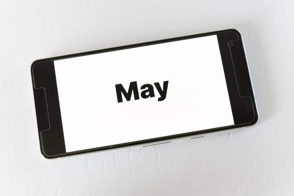 Black smartphone lying at an angle on a white textured surface, displaying the word "May" in bold black text on a white screen.