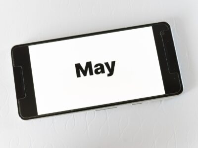Black smartphone lying at an angle on a white textured surface, displaying the word "May" in bold black text on a white screen.