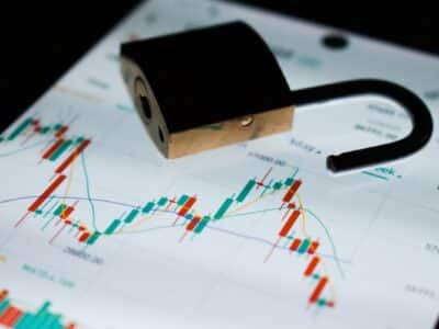 Open brass and black padlock resting on a screen displaying multicolored candlestick charts, line graphs and bar charts with numerical values.