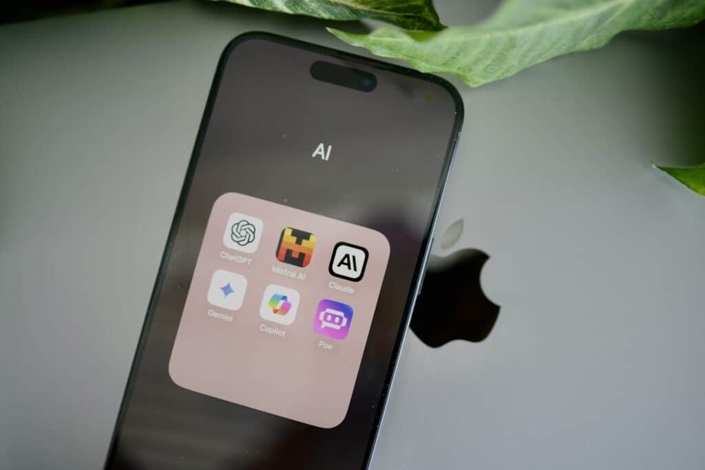 Close-up of an iPhone screen displaying an open app folder labeled "AI" containing six app icons — ChatGPT, Mistral AI, Claude, Gemini, Copilot, and Poe — with the Apple logo visible on the back of a laptop and a green leaf at the top of the frame.