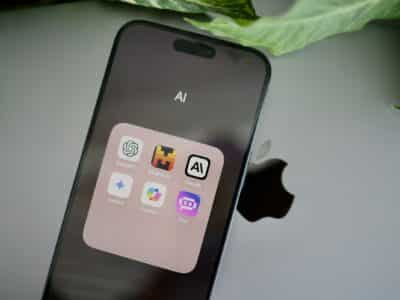 Close-up of an iPhone screen displaying an open app folder labeled "AI" containing six app icons — ChatGPT, Mistral AI, Claude, Gemini, Copilot, and Poe — with the Apple logo visible on the back of a laptop and a green leaf at the top of the frame.