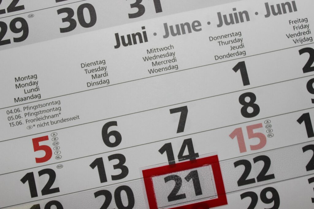 Close-up of a multilingual wall calendar page for June, with weekday headers in German, English, French and Dutch. The dates 12 through 22 are visible across two rows, with the number 21 highlighted by a red rectangular marker overlay. The numbers 5 and 15 appear in red text with country code annotations in small circles next to them.