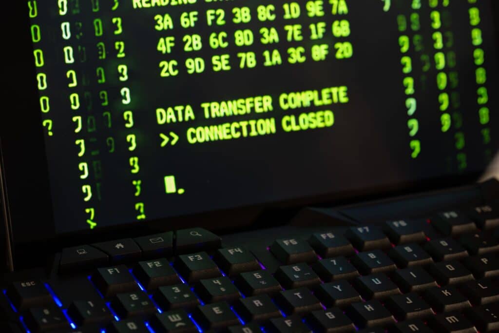 Close-up of a backlit black keyboard with red and blue key lighting in front of a monitor displaying rows of green hexadecimal values and the text "DATA TRANSFER COMPLETE >> CONNECTION CLOSED."