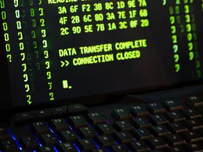 Close-up of a backlit black keyboard with red and blue key lighting in front of a monitor displaying rows of green hexadecimal values and the text "DATA TRANSFER COMPLETE >> CONNECTION CLOSED."