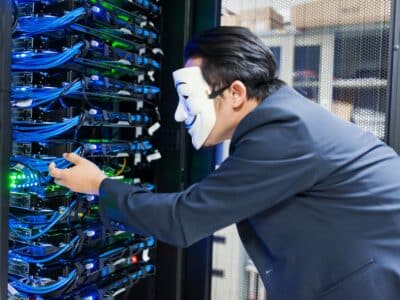 Caucasian male in a grey business suit and Guy Fawkes mask reaching into a lit, blue server rack inside a data center as he unplugs a wire.