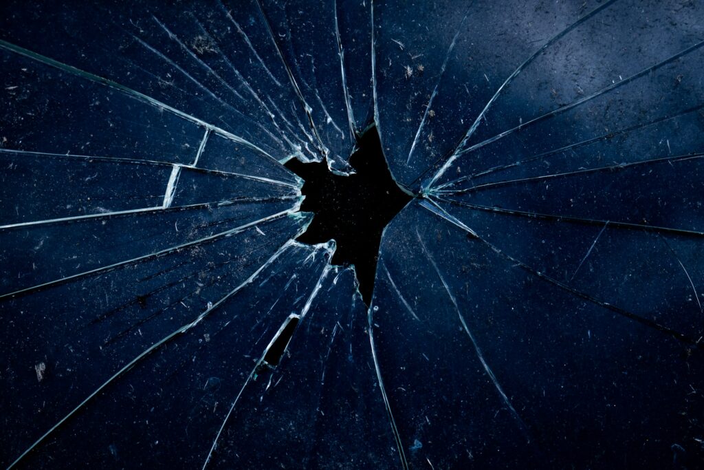 Close-up of dark blue-tinted glass shattered around a small central impact hole, with fractures radiating outward in a spider-web pattern against a black background.