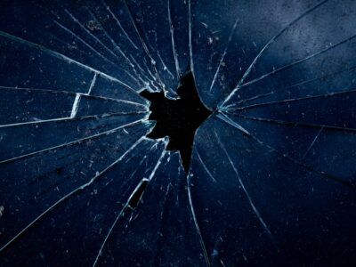 Close-up of dark blue-tinted glass shattered around a small central impact hole, with fractures radiating outward in a spider-web pattern against a black background.