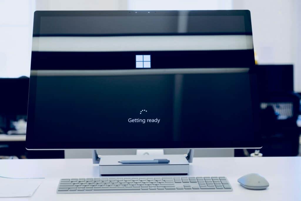 Microsoft Surface Studio all-in-one desktop displaying a Windows setup screen — a large Windows logo above the text "Getting ready" and a small circular loading indicator — with a Surface Pen resting on the base of the stand, a Surface keyboard and Surface Mouse arranged on the white desk in front and a softly blurred office in the background.