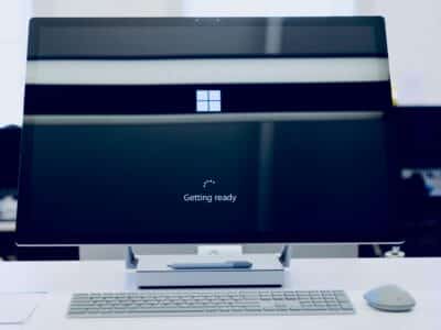 Microsoft Surface Studio all-in-one desktop displaying a Windows setup screen — a large Windows logo above the text "Getting ready" and a small circular loading indicator — with a Surface Pen resting on the base of the stand, a Surface keyboard and Surface Mouse arranged on the white desk in front and a softly blurred office in the background.