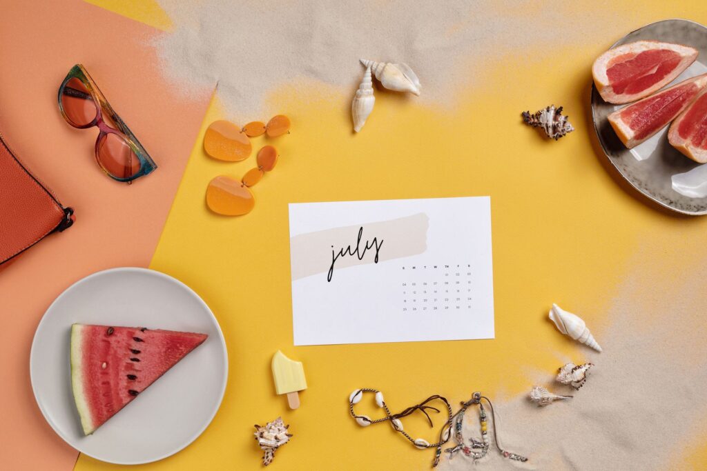 Flat lay of a paper July calendar on a yellow and coral background surrounded by rainbow-tinted sunglasses, a coral leather bag, orange drop earrings, assorted seashells, a watermelon wedge on a white plate, grapefruit slices on a gray plate and shell bracelets.