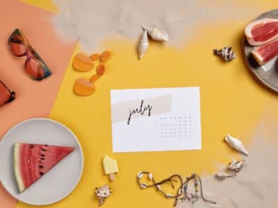 Flat lay of a paper July calendar on a yellow and coral background surrounded by rainbow-tinted sunglasses, a coral leather bag, orange drop earrings, assorted seashells, a watermelon wedge on a white plate, grapefruit slices on a gray plate and shell bracelets.