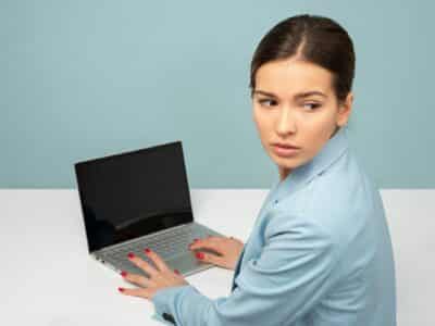 Woman in a light blue blazer seated at a white desk with both hands resting on a silver laptop keyboard, turning to look back over her shoulder toward the camera with a guarded expression, the dark laptop screen angled away from view against a pale blue-green background.