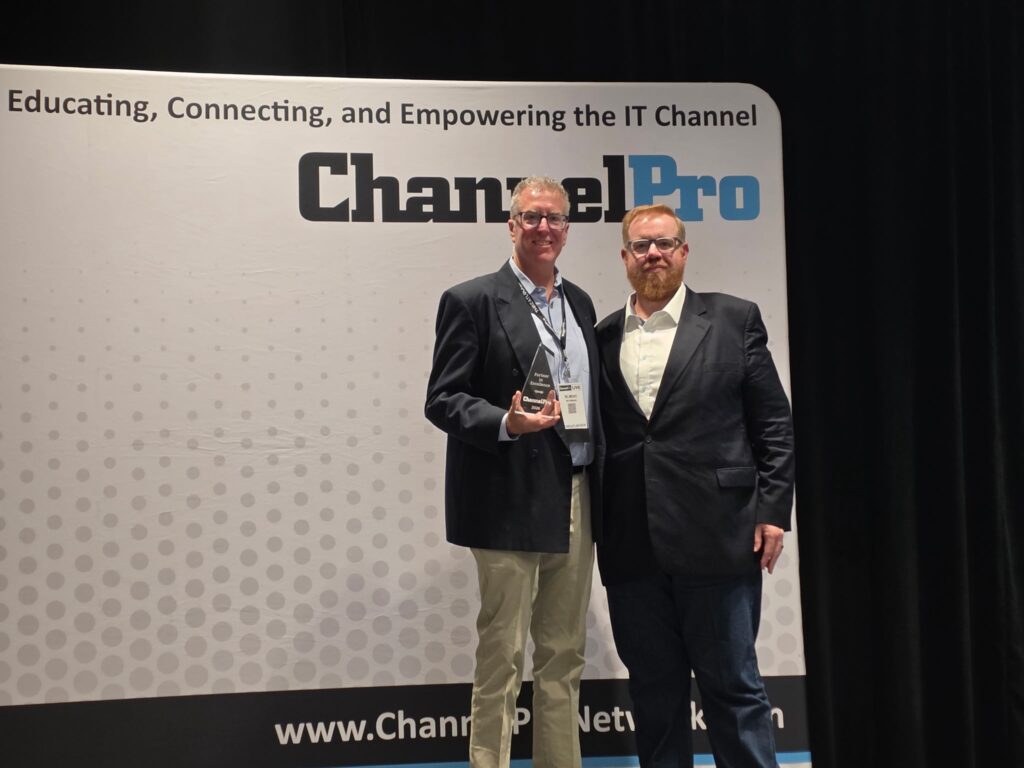 Two Caucasian men in blazers stand in front of a ChannelPro backdrop, while the one on the left holds a glass Partner in Excellence trophy.