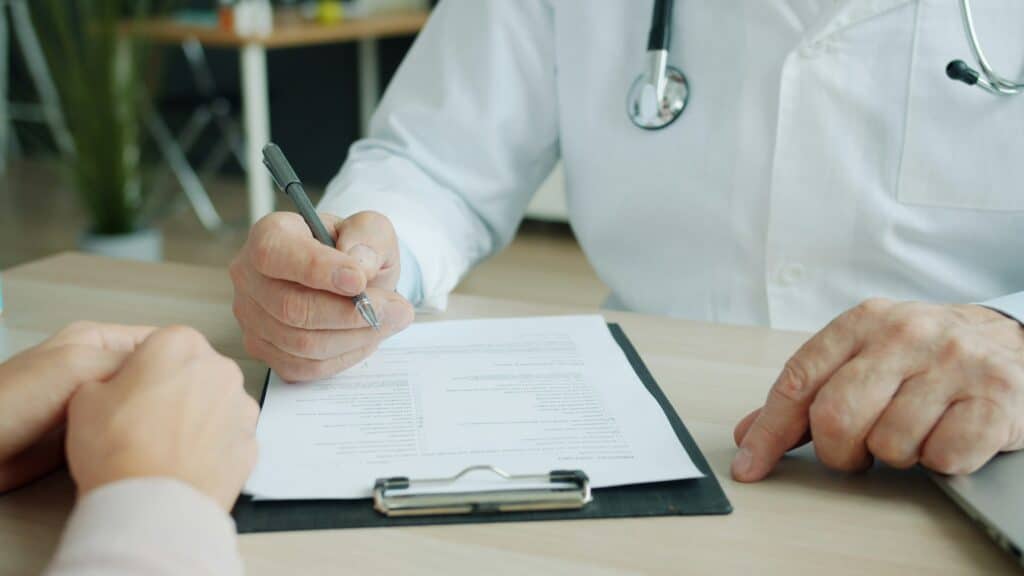Cropped view of a person in a white coat with a stethoscope holding a pen over a printed medical history form on a black clipboard, with another person's hands resting on the wooden desk opposite.