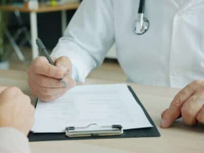 Cropped view of a person in a white coat with a stethoscope holding a pen over a printed medical history form on a black clipboard, with another person's hands resting on the wooden desk opposite.