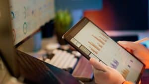 Close-up of a person's hands holding a tablet displaying bar and line chart dashboards, with a stylus, an open laptop keyboard and a printed board of percentage dials blurred behind.
