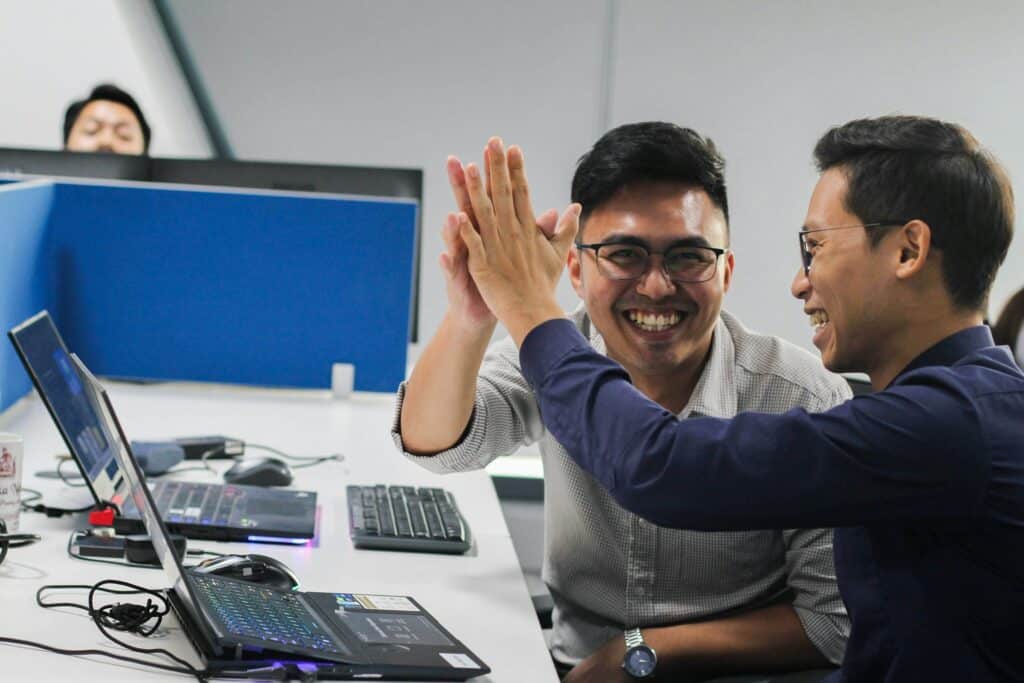 Two men in an office high-five over a desk with two laptops, a keyboard, and a mouse, with a third man seated at a monitor in the background.