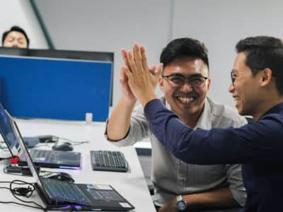 Two men in an office high-five over a desk with two laptops, a keyboard, and a mouse, with a third man seated at a monitor in the background.