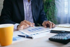 Person in a navy blazer and blue plaid shirt, face out of frame, touching a white tablet showing blue bar charts, with printed charts, a black pen, a calculator, a ring-bound notebook and a yellow mug on a wooden desk.