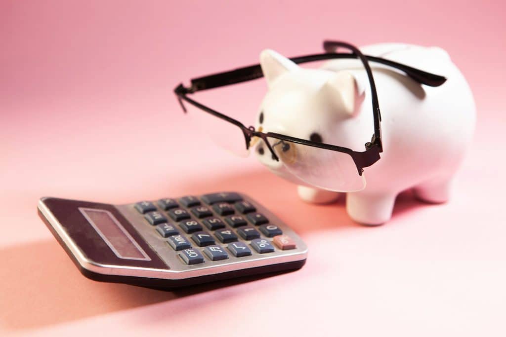 White ceramic piggy bank with a pinkish hue and wearing oversized black half-rim glasses resting on its snout, facing a black and gray desktop calculator on a pink background, representing IT budget planning for 2027.