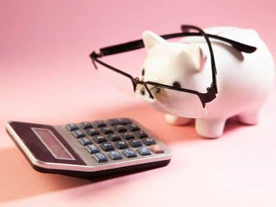 White ceramic piggy bank with a pinkish hue and wearing oversized black half-rim glasses resting on its snout, facing a black and gray desktop calculator on a pink background, representing IT budget planning for 2027.